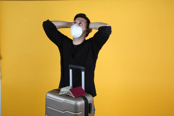 man with protective mask suitcase and passport, listening to music, his flight is delayed by covid-19, yellow background - Photo, Image