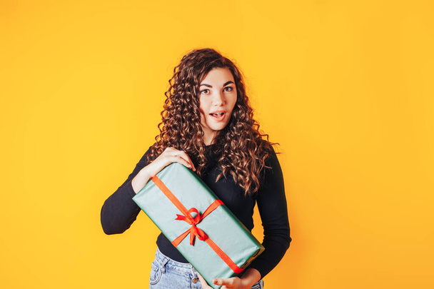 Schöne Frau in einer schwarzen Jacke auf gelbem Hintergrund, die eine große Schachtel mit einem Geschenk in ihren Händen hält. - Foto, Bild