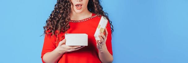 Beautiful woman in a red dress on a blue background opens a box with a gift for the holiday. - Foto, afbeelding