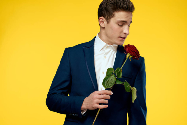Mladý muž v klasickém obleku s červenou růží v ruce na žlutém pozadí emoce oříznuté pohled modelu - Fotografie, Obrázek