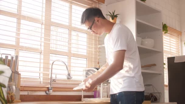 Gelukkige jonge Aziaat die de afwas doet terwijl hij schoonmaakt in de keuken. Thuis blijven, zelf isoleren, sociale afstand nemen, quarantaine voor coronavirus, lifestyle man thuis concept. - Video