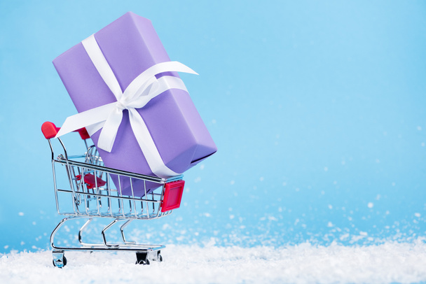 Little gift box in shopping trolley and artificial snow on blue background, new year concept - Foto, Imagem