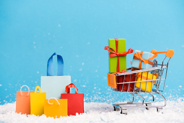Tiny paper bags near trolley with bunch of little gifts on blue background, new year concept - Photo, Image