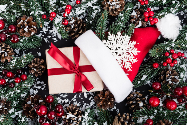 Yapay karlı ladin dalları, Noel Baba şapkası yanında hediye kutusu ve siyah arka planda dekoratif kar tanesi. - Fotoğraf, Görsel