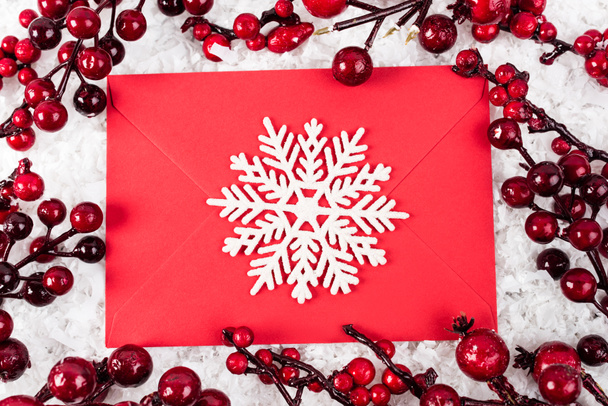 Bovenaanzicht van decoratieve sneeuwvlok op rode envelop bij takken met kunstmatige bessen op witte textuur achtergrond, nieuwjaarsconcept - Foto, afbeelding