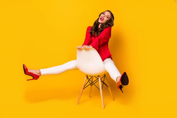 Full length photo of attractive business lady sit cosy chair between legs cheerful playful mood wear red shirt white trousers high-heels isolated yellow bright color background - Фото, зображення