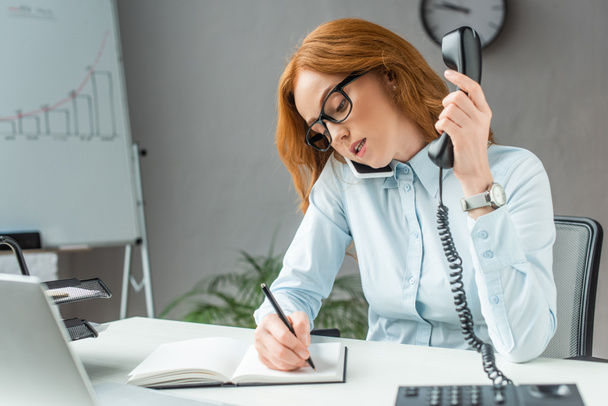 Femme d'affaires rousse avec combiné parlant sur un téléphone mobile, tout en écrivant dans un carnet sur le lieu de travail sur fond flou - Photo, image