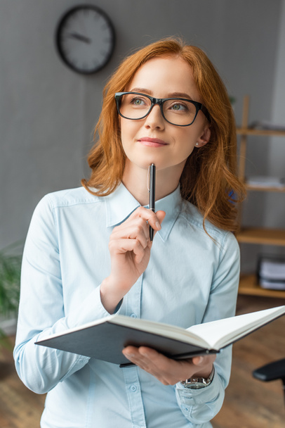Positive businesswoman with notebook thinking, while looking away with blurred office on background - Photo, Image
