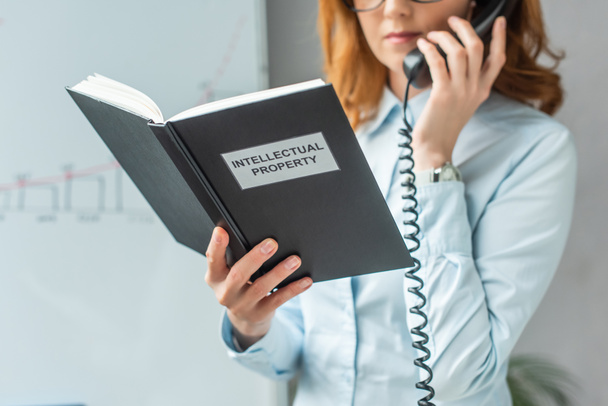 Gewassen beeld van zakenvrouw met een boek met intellectuele eigendom belettering, terwijl het praten op vaste telefoon op wazige achtergrond - Foto, afbeelding
