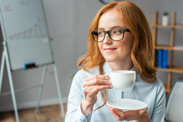 Femme d'affaires rousse souriante regardant ailleurs, tout en tenant une tasse de café et une soucoupe avec un tableau à feuilles floues sur fond - Photo, image
