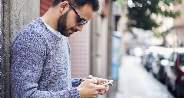 Side view of young bearded male wearing warm knitted sweater and sunglasses browsing smartphone while standing on street in daytime - Фото, изображение
