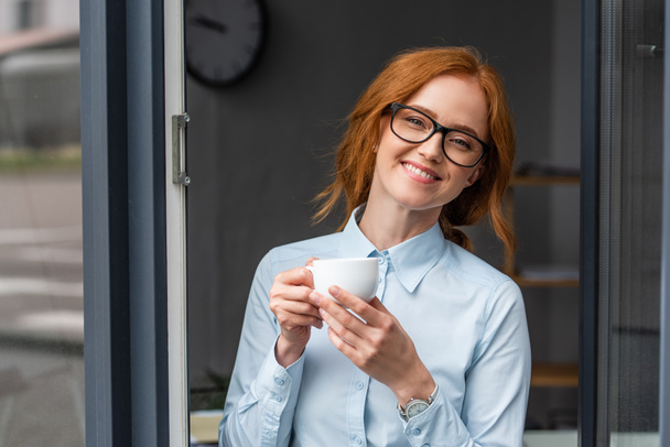 Heureuse femme d'affaires rousse avec tasse de café regardant la caméra à la fenêtre - Photo, image