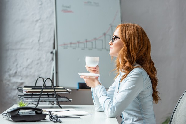 İş kadını, fincan tabağı ve kahve fincanı ile yan gözle bakarken, iş yerinde bulanık arka planda otururken, - Fotoğraf, Görsel