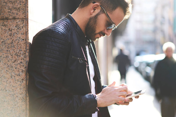 Crop side view of neat man with beard in black glasses standing near building leaning on wall looking down at screen of mobile phone and messaging - Foto, afbeelding