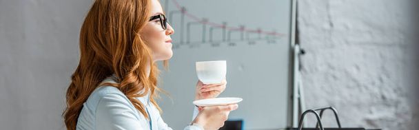 Vista lateral de la mujer de negocios pelirroja con taza de café y platillo mirando hacia otro lado con el rotafolio borroso en el fondo, pancarta - Foto, Imagen