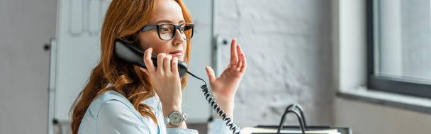 Redhead businesswoman gesturing while talking on landline telephone with blurred flipchart on background, banner - Foto, imagen