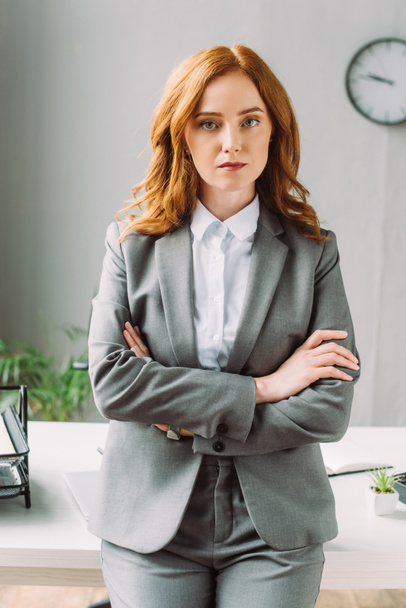 Front view of serious businesswoman with crossed arms looking at camera near workplace on blurred background - Foto, afbeelding
