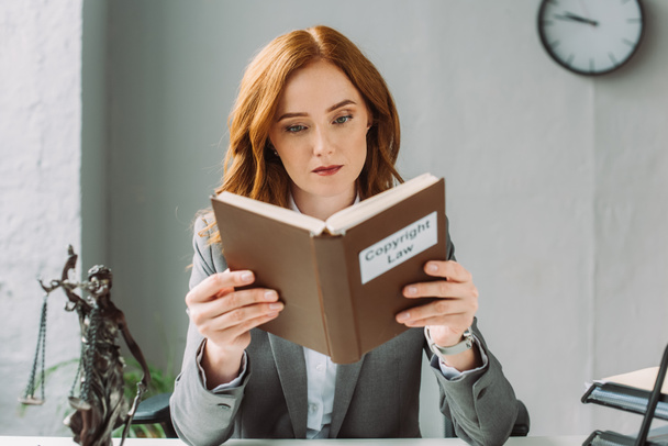 Сосредоточенная женщина-юрист читает книгу с надписью авторского права рядом с фигуркой в офисе на размытом фоне - Фото, изображение