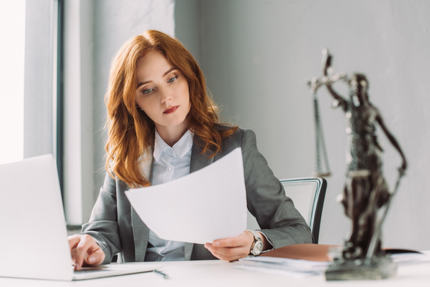 Redhead lawyer looking at paper sheet, while sitting near laptop at table with blurred themis figurine on foreground - Фото, зображення