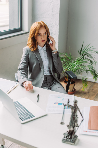 Abogada confiada hablando por teléfono móvil, mientras está sentada cerca del lugar de trabajo con la figura borrosa en primer plano - Foto, Imagen