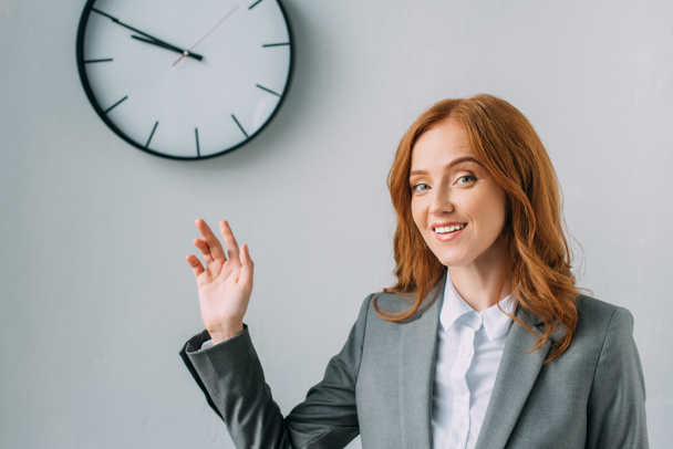 Smiling businesswoman in formal wear pointing with hand at wall clock on grey - Photo, Image