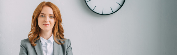 Positive redhead businesswoman looking at camera near wall clock on grey, banner - Фото, изображение