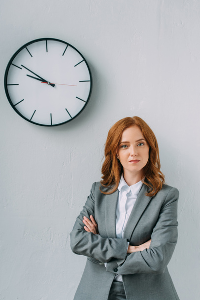Front view of confident businesswoman with crossed arms looking at camera near wall clock on grey  - Foto, Imagem