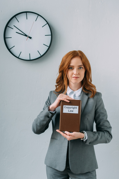Front view of business woman looking at camera, while showing book with copyright law lettering near wall clock on grey - Фото, изображение