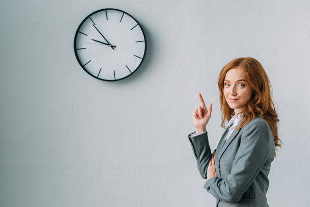 Smiling redhead businesswoman pointing with finger at wall clock, while looking at camera on grey - Fotografie, Obrázek