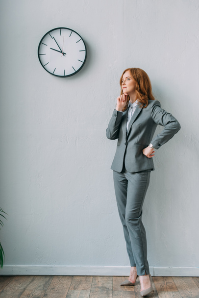 Volledige lengte van doordachte zakenvrouw met de hand op hip staan in de buurt muur klok in kantoor - Foto, afbeelding