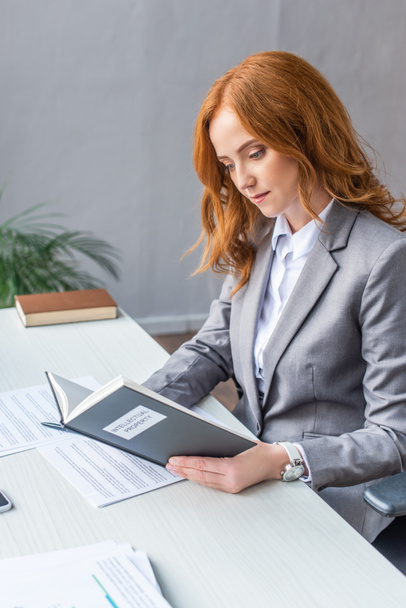 Redhead lawyer reading book with intellectual property lettering at workplace with documents on blurred background - Foto, Imagen