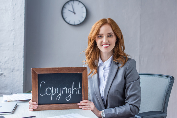 Mosolygó női ügyvéd néz kamera, miközben mutatja tábla szerzői jogi felirat a munkahelyen elmosódott háttér - Fotó, kép