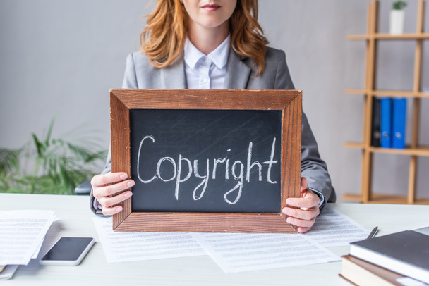 Ausgeschnittene Ansicht eines Rechtsanwalts, der eine Tafel mit dem Schriftzug "Urheberrecht" in der Hand hält, während er mit Dokumenten auf verschwommenem Hintergrund am Tisch sitzt - Foto, Bild