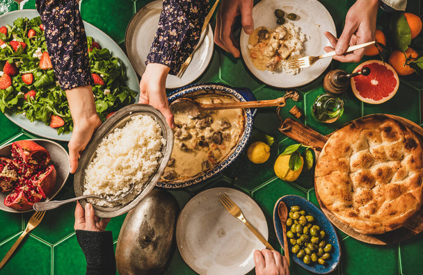 Una cena turca in famiglia. Piatto di persone che passano riso pilav sulla tavola verde con agnello in salsa di yogurt, rucola fresca e insalata di fragole e focaccia, vista dall'alto. Cena di iftar di Ramazan - Foto, immagini