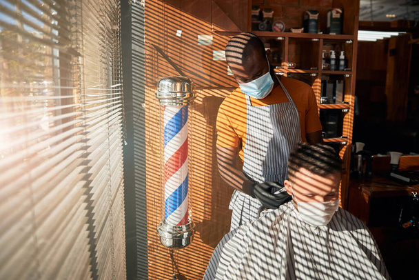 Tıp maskeli berber berber dükkanındaki adama saç kesimi yapıyor. - Fotoğraf, Görsel