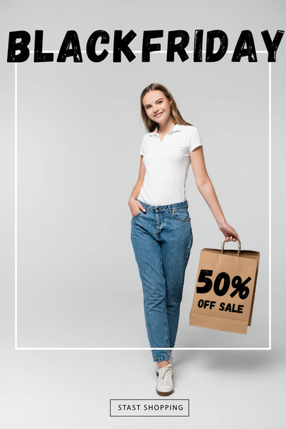 joyful young woman holding shopping bag while standing with hand in pocket near black friday and 50 percent off sale lettering on grey - Photo, Image