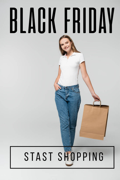 joyful young woman holding shopping bag while standing with hand in pocket near black friday lettering on grey - Photo, Image