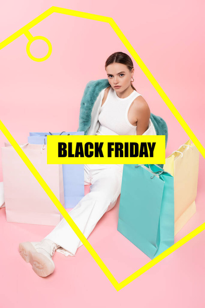 high angle view of young woman in blue faux fur jacket sitting near shopping bags and black friday lettering and tag illustration on pink - Foto, imagen