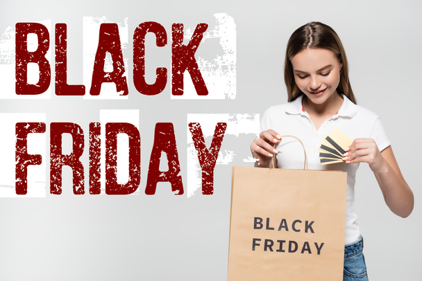joyful woman holding credit cards and looking at shopping bag near black friday lettering on grey - Photo, Image