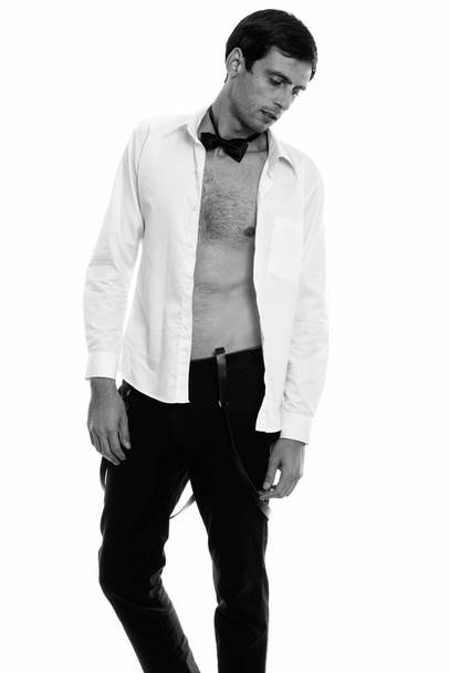 Studio shot of young handsome man thinking and looking down with open shirt while wearing bow tie - Foto, imagen