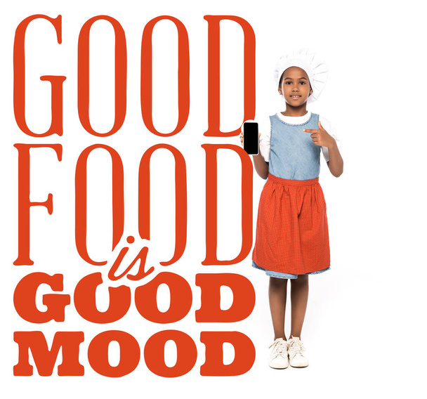 niño afroamericano vestido con traje de chef señalando con el dedo en el teléfono inteligente cerca de la buena comida es letras de buen humor en blanco  - Foto, Imagen