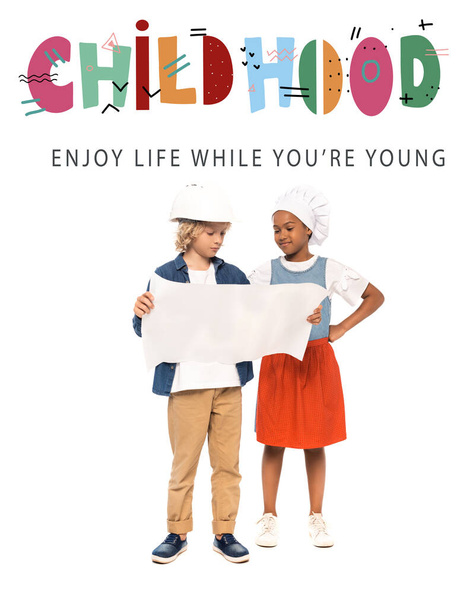 curly boy in safety helmet and american kid in chef hat looking at blueprint near childhood, enjoy life while you are young lettering on white  - Photo, Image