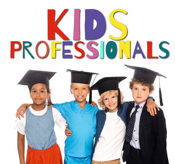 multikulturelle Kinder mit Abschlussmützen in Kostümen verschiedener Berufe in der Nähe von Kids Professionals Schriftzug auf weiß  - Foto, Bild