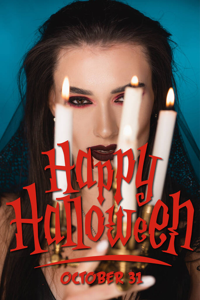 young woman in veil with blood on face near burning candles and happy halloween lettering on blue - Фото, изображение