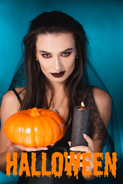 pale woman with black makeup, veil holding burning candle and pumpkin near halloween lettering on blue - Foto, Imagen
