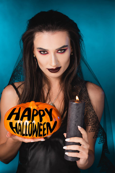 pale woman with black makeup and veil holding burning candle and pumpkin near happy halloween lettering on blue - Fotoğraf, Görsel