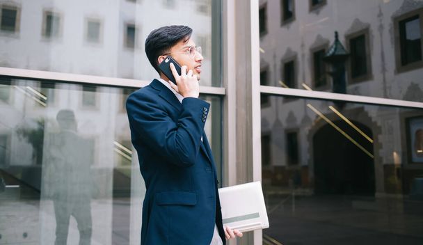 Side view of young male in formal wear and eyeglasses talking on mobile phone while standing near glass wall of modern building - Photo, image