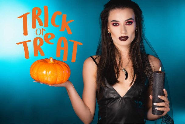 young pale woman with black makeup and veil holding candle and pumpkin near trick or treat on blue - Zdjęcie, obraz