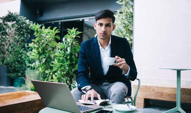 Confident handsome 20s businessman in trendy wear looking at camera holding aroma coffee working remotely, serious hindu proud ceo using laptop computer and 4G internet connection for freelance job - Foto, immagini