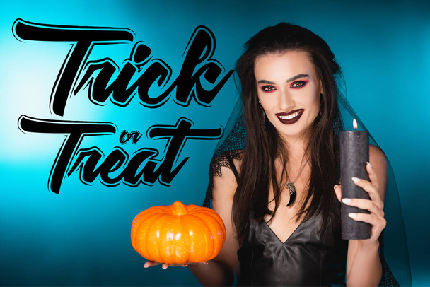 joyful woman with black makeup and veil holding burning candle and pumpkin near trick or treat lettering on blue - Photo, Image